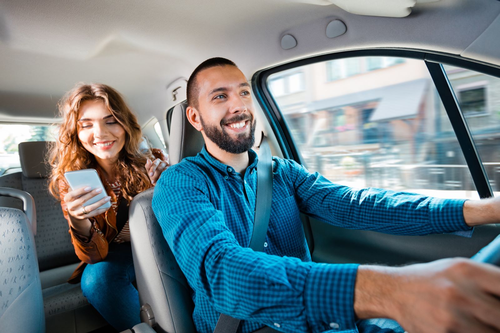 A person looking at their phone in the backseat of a car with a person directly in front of them in the driver's seat driving the car.