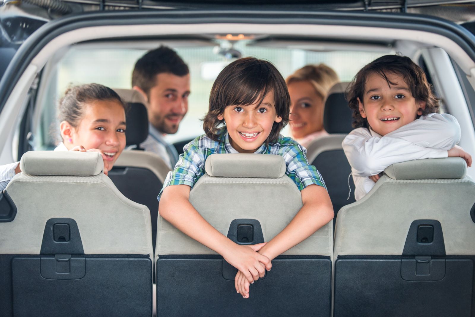 Three children looking out the back side of a car. Two adults are in the front seats of the car.