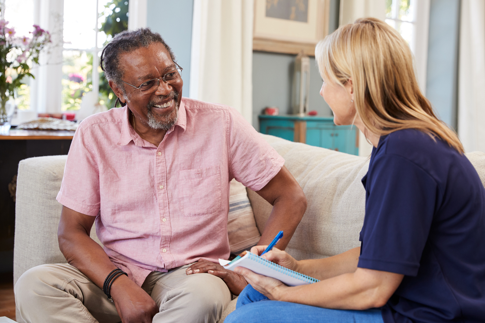 An older adult man sits on a sofa with an aide who is holding a clipboard with forms.