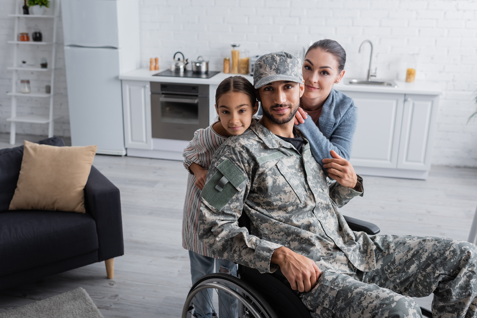 Dressed in military uniform, a soldier sits in a wheelchair while being embraced by his wife and daughter.