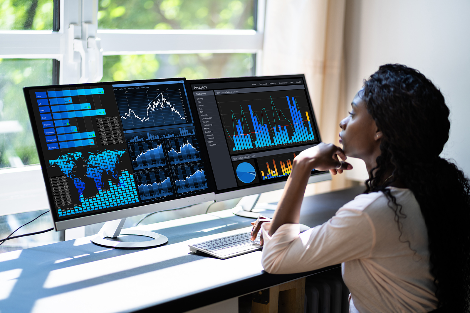 A Black woman analyzes data using a computer. She has long, braided hair and wears business casual clothing. Her desk is in a sunny office.