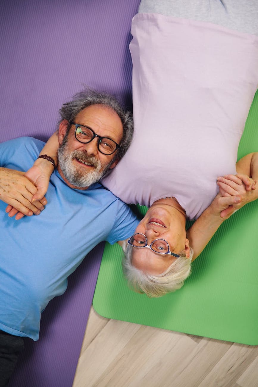 An independent older couple lays on yoga mats and embraces. The man has gray hair, a beard and eyeglasses. The woman has short white hair, glasses and wears a wristwatch.