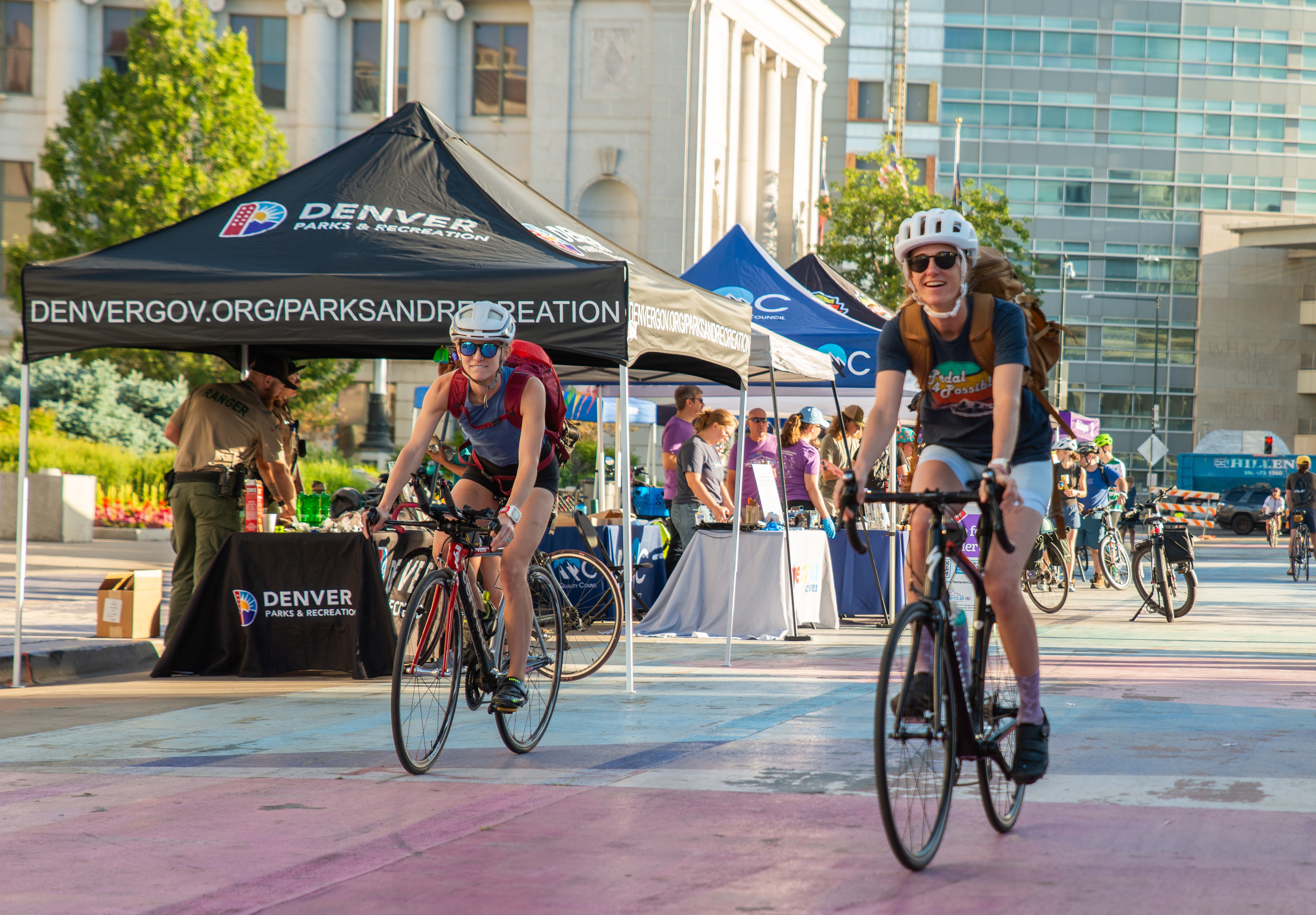 Two people riding bicycles with a crowd of people and booths set up in the background. This image was taken on Bike to Work Day 2024.