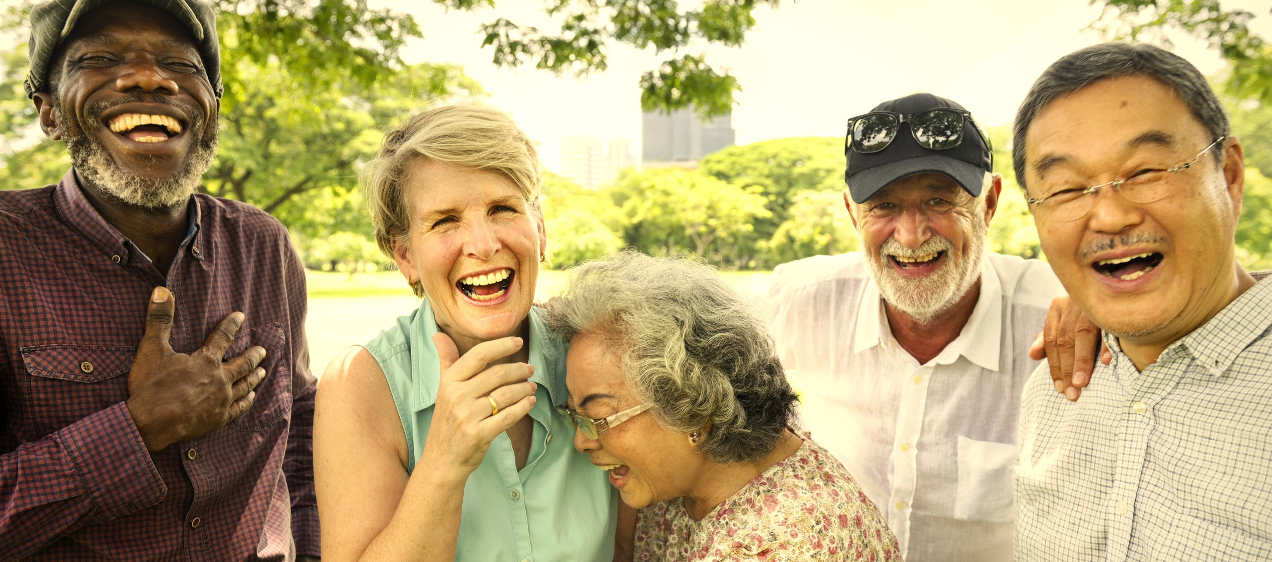 Four older adults,  a Black man, a white woman, an Asian woman, a white man and an Asian man are standing while laughing in a park setting.