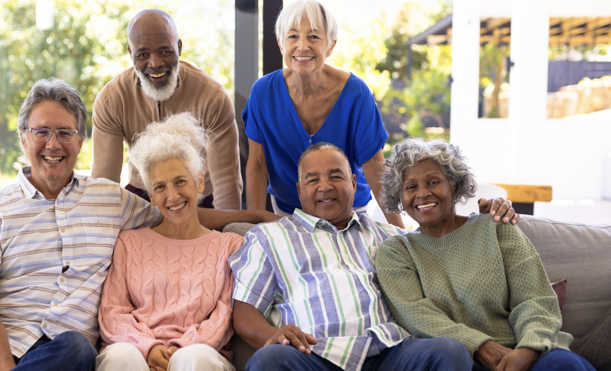 A racially diverse group of four older adults of various genders sit on a couch. Two older adults stand behind the couch.