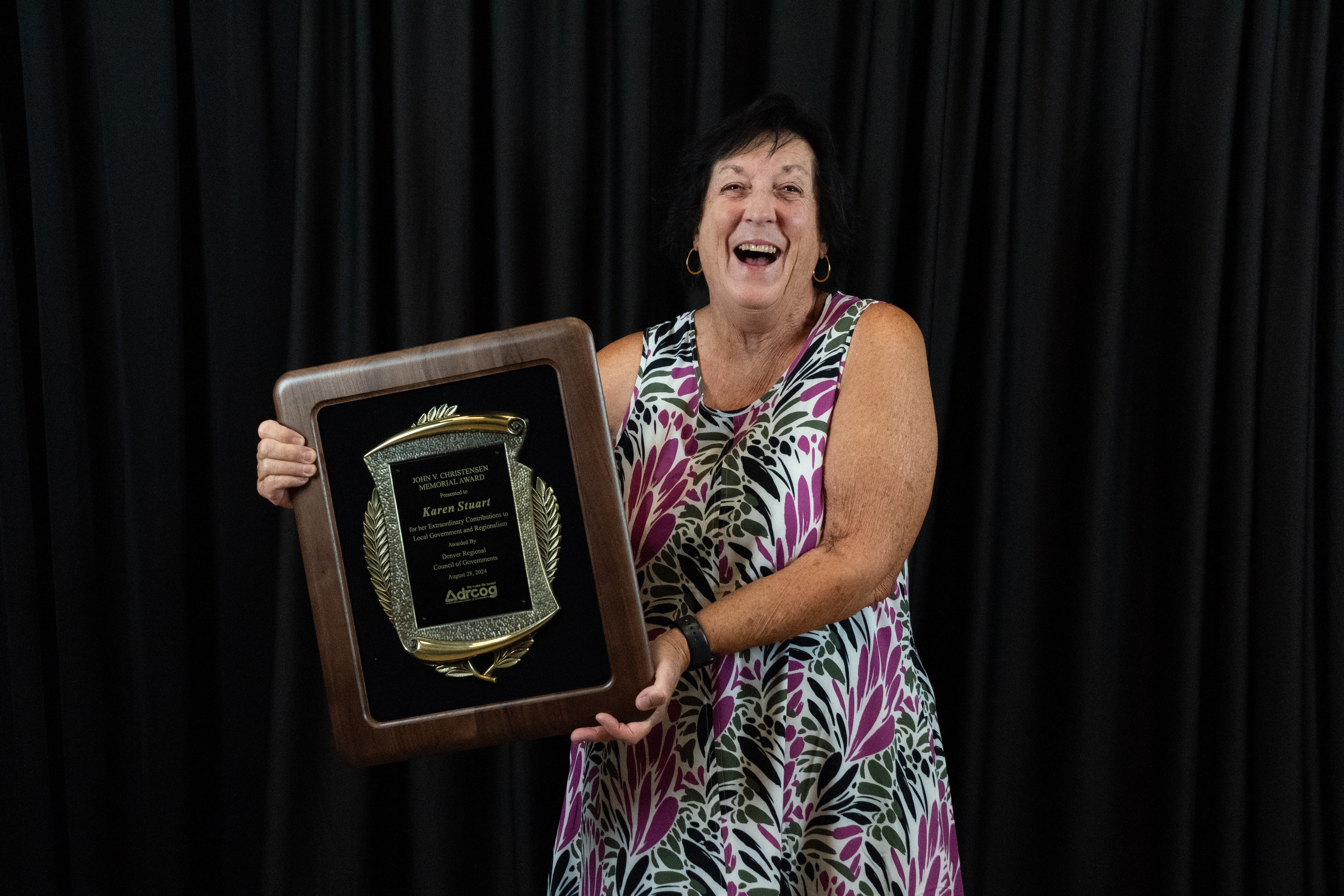 A middle-aged white woman with a joyous smile and colorful dress holds a large engraved plaque.