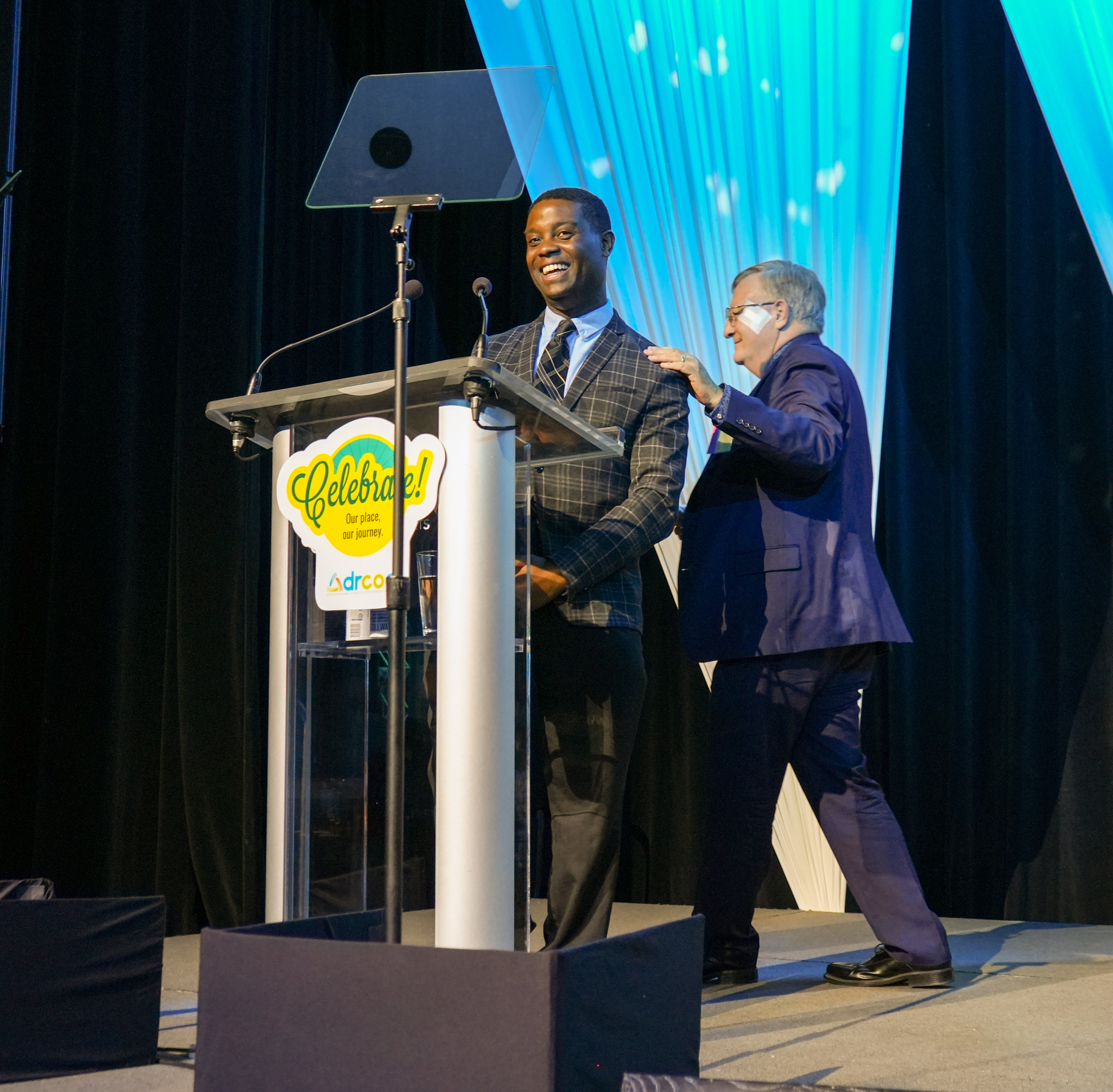 An older white man touches the shoulder of a younger Black man at a lectern as he walks behind him.