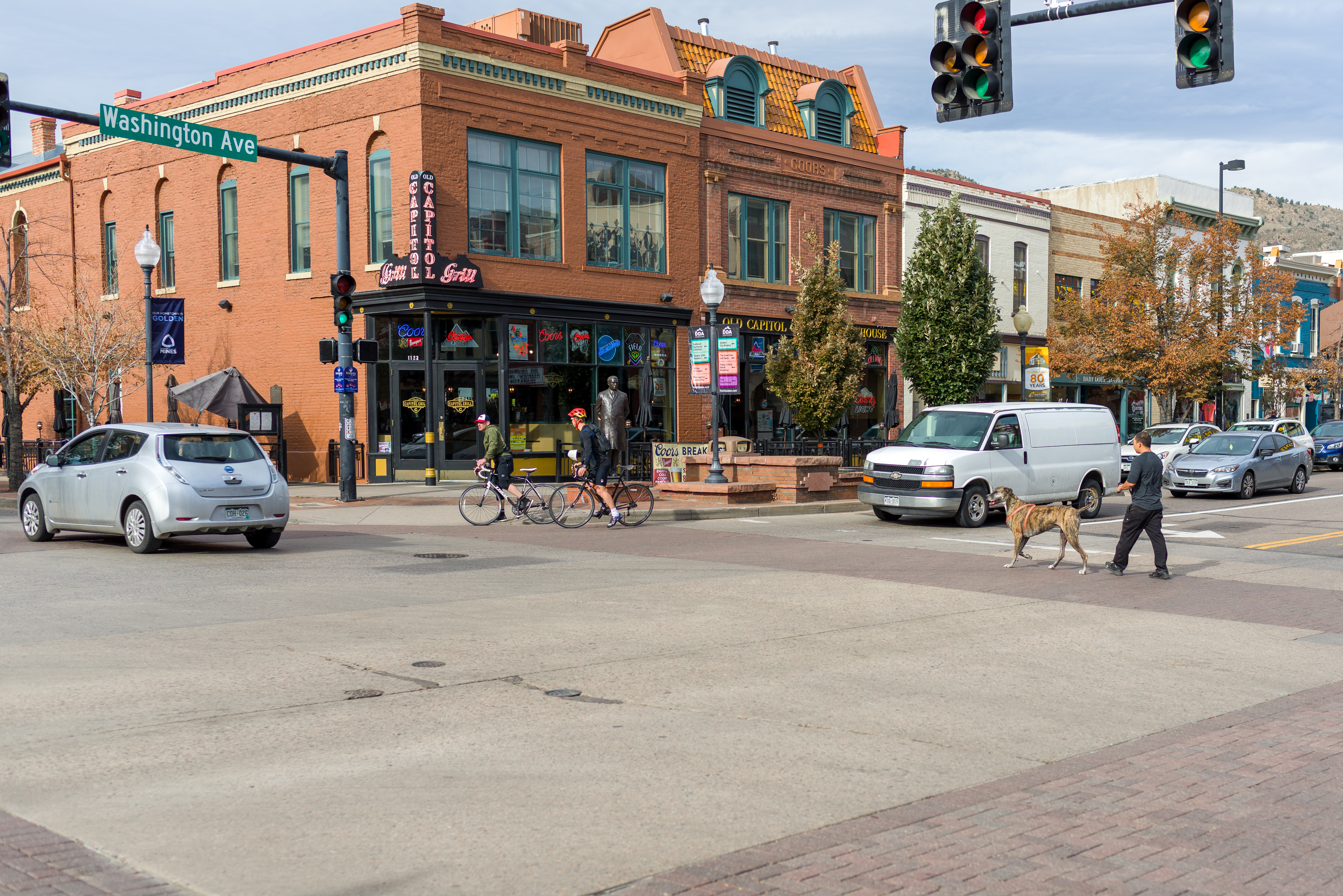 Downtown Golden, Colorado, two people are riding bicycles as a silver car passes them and a man walks a dog through an intersection as cars are stopped.